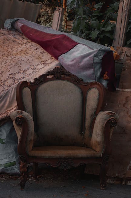 Inside a property, an ornate vintage armchair with a high carved wooden frame and light upholstery is positioned in front of a partially visible wooden table covered with a patterned fabric. Behind the armchair, a large rolled-up piece of fabric, possibly a rug or drapery, is placed on top of the table, with its edges hanging over. To the right, part of a door or window frame and some green foliage are visible, indicating an outdoor access point. The scene suggests an ongoing home relocation or furniture removal process, with the furniture and materials arranged for packing or transportation. Natural lighting reveals the details of the furniture and objects, reflecting a typical environment during a house move, with items being prepared for loading onto a removal vehicle, possibly aligning with professional removal services by [COMPANY_NAME] for house removals in Notting Hill.