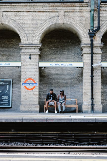 Two young individuals sit on a wooden bench at the Notting Hill Gate London Underground station platform, positioned against a backdrop of historic brickwork with large stone archways. The station's blue and white signage, including the 'NOTTING HILL GATE' sign and the London Underground roundel, is visible above them. The platform edge is in the foreground, with train tracks running parallel, and a small poster displayed on a wall to the left. This scene depicts typical public transport waiting area surroundings in an urban setting. During a home relocation or furniture transport process, nearby removal services like those at removalcompaniesnottinghill.co.uk may assist with moving bulky furniture, as evidenced by the potential for such objects being loaded into or transported from this station area. The overall environment is well-lit, with the brick façade and transport signage clearly visible, reflecting the busy yet organized atmosphere of a central London train station used during packing and moving logistics.