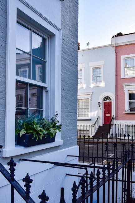 A close-up view of a white-painted brick house facade in a residential area, featuring a large double-hung sash window with a white frame and a window box filled with green plants and flowers. The window is partially open, revealing interior curtains. In the foreground, black metal railings enclose a small front step area. Adjacent houses are visible, including a pastel pink building with white window frames and a dark grey roof. The scene is well-lit with natural daylight, and the environment suggests a quiet, urban street setting suitable for home relocation and furniture transport activities, as seen in professional removals involving packing and loading processes.