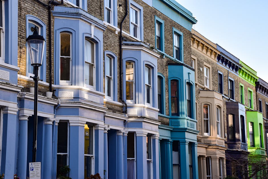A row of multi-storey Victorian terraced houses in Notting Hill painted in vibrant pastel colours, including light blue, turquoise, cream, black, and lime green. The buildings feature bay windows with white trims, decorative cornices, and arched window frames. A black street lamp stands in the foreground on the pavement, which is partially visible with a parked car and a parking sign. The scene is set during daylight with clear weather, highlighting the contrast of the colourful facades. This image reflects urban residential architecture typical of home relocation and furniture transport services in Notting Hill, with an emphasis on building exteriors, window details, and the street environment where house removals might take place.