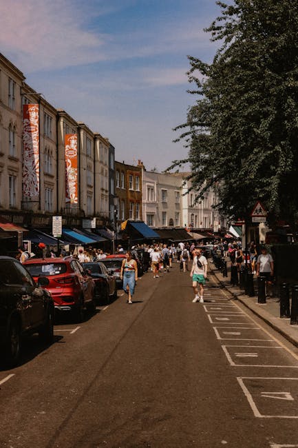 A busy street scene on Portobello Road in Notting Hill, with pedestrians walking along the pavement and some carrying boxes and small luggage items, indicating a home relocation or moving process. Several parked cars line the curb on both sides of the road, with storefronts and residential buildings visible in the background. On the right, a large leafy tree partially shades the sidewalk, and a traffic warning sign is seen further down the street. The presence of people and the layout suggest a vibrant area where careful planning for removals and furniture transport may be required, aligning with services such as house removals provided by [COMPANY_NAME], especially when considering moving logistics and packing and moving needs. The scene is illuminated by natural daylight, with a clear sky overhead, emphasizing the urban environment typical of a busy residential district in Notting Hill.
