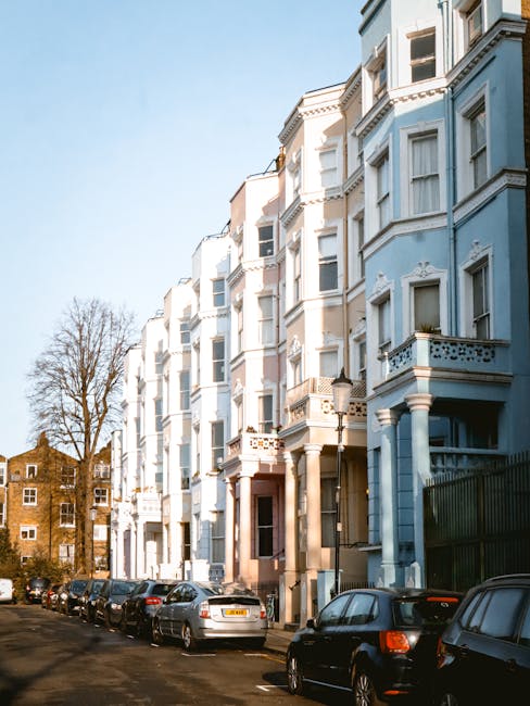 Photograph of a row of multi-storey residential buildings in Notting Hill, painted in shades of light blue and cream, with classic architectural details such as bay windows, decorative moldings, and balconies. The street is lined with parked cars and a narrow pavement runs along the front of the buildings. In the foreground, a removal company’s van is parked, partially visible on the right, with the side door open and a few packed cardboard boxes, wrapped in plastic and fabric covers, are being loaded inside or onto a trolley nearby, suggesting a home relocation or moving process. A few large furniture items, possibly a wardrobe or cabinet, are visible among the packing materials. The scene is set during daylight with clear skies, and in the background, trees and additional residential structures are visible, emphasizing an urban environment familiar for house removals and furniture transport in the area near Portobello Road. This image illustrates typical elements involved in packing and loading during moving services provided by companies like [COMPANY_NAME].
