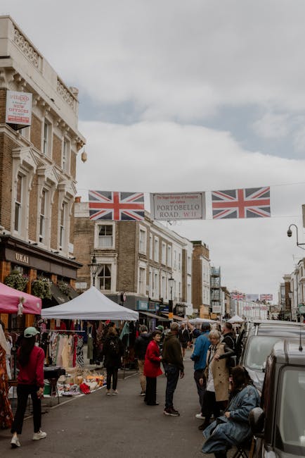A busy street scene on Portobello Road in Notting Hill, with pedestrians walking along the pavement and some carrying boxes and small luggage items, indicating a home relocation or moving process. Several parked cars line the curb on both sides of the road, with storefronts and residential buildings visible in the background. On the right, a large leafy tree partially shades the sidewalk, and a traffic warning sign is seen further down the street. The presence of people and the layout suggest a vibrant area where careful planning for removals and furniture transport may be required, aligning with services such as house removals provided by [COMPANY_NAME], especially when considering moving logistics and packing and moving needs. The scene is illuminated by natural daylight, with a clear sky overhead, emphasizing the urban environment typical of a busy residential district in Notting Hill.