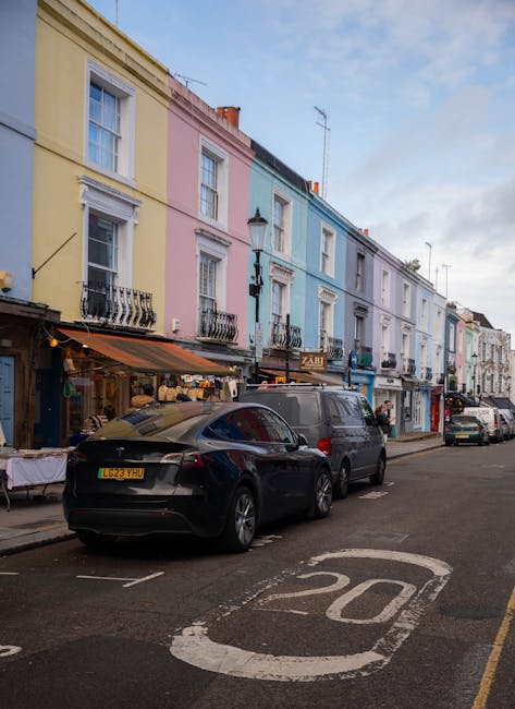 A white residential building on Portobello Road displaying a street sign for Portobello Road, W.11, attached to the exterior wall. The building features a small front porch with a decorative white balustrade and two columns supporting the ceiling. In the foreground, there are cardboard boxes and packing materials placed on the pavement, indicating packing preparations for a home relocation. The beige cardboard boxes vary in size and are stacked or arranged for transport. Behind the boxes, a black moving trolley with handles is visible, along with a portion of a black vehicle, possibly a removal van, parked in the loading area. The surroundings include some greenery with trees and bushes, and soft daylight illuminates the scene. The setting captures the typical logistics involved in furniture transport and packing during a house move, illustrating the careful handling of items for a professional removal service in Notting Hill, such as those offered by [COMPANY_NAME] as part of their removals service category.
