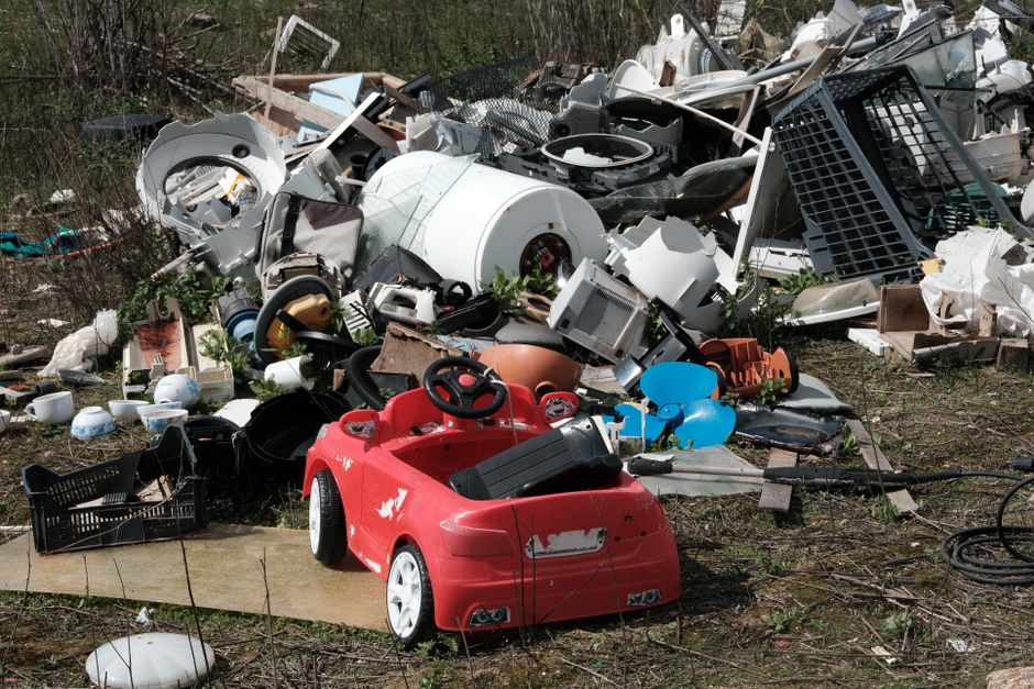 A large pile of household waste and discarded items outdoors, including broken appliances, plastic containers, and various household objects scattered across a grassy area. In the foreground, a red children's toy car with visible scratches and worn paint sits on a piece of cardboard. Behind the toy, there are numerous items such as washing machines, broken furniture, plastic bins, and cardboard boxes, some of which are partially crushed or damaged. The pile extends onto a paved area, with Mr. and Mrs. removal companies Notting Hill visible in the background, and a few gardening bushes and trees surrounding the scene. The objects are arranged in a disorganized manner, illustrating waste disposal after a home move or renovation, highlighting the importance of proper household waste management during house removals and furniture transport processes. The lighting appears natural, indicating daytime.