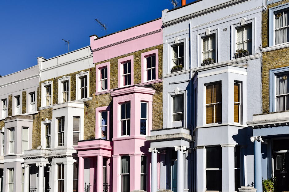A row of multi-storey terraced houses with Victorian-style architecture, featuring large sash windows, decorative cornices, and bay windows on the ground and first floors. The buildings are brickwork with some facades painted in pastel colours, including white and light pink. The image shows a clear blue sky, with sunlight casting shadows on the building fronts. The areas below the windows and the bay windows on some sections have potted plants, and the facades are clean and well-maintained. This residential street scene is relevant to house removals and moving services, illustrating typical urban home exteriors encountered during home relocation or furniture transport processes, as provided by companies like [COMPANY_NAME].