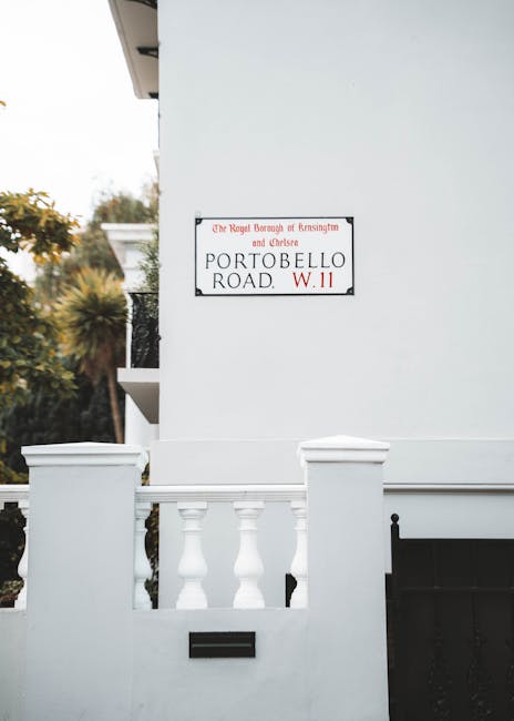 An exterior view of a white residential building on Portobello Road in Notting Hill, showing a section of the front porch with white balustrades and a small mailbox attached to the wall beneath a street sign that reads 'Kensington & Chelsea Council rules for Notting Hill moves,' indicating local regulations for house removals. The image captures the upper part of the building with a partial view of the overhanging roof, and in the background, there are trees with green and orange foliage suggesting a garden or outdoor space. The scene is well-lit with natural daylight, emphasizing the clean, painted surfaces of the house exterior and the neatly maintained architectural details. This setting is relevant to moving and packing services by illustrating the typical facade where furniture transport and home relocation activities take place, aligned with professional removals and adherence to local council rules.