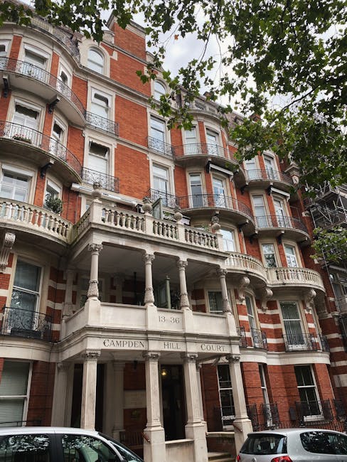 An exterior view of a white residential building on Portobello Road in Notting Hill, showing a section of the front porch with white balustrades and a small mailbox attached to the wall beneath a street sign that reads 'Kensington & Chelsea Council rules for Notting Hill moves,' indicating local regulations for house removals. The image captures the upper part of the building with a partial view of the overhanging roof, and in the background, there are trees with green and orange foliage suggesting a garden or outdoor space. The scene is well-lit with natural daylight, emphasizing the clean, painted surfaces of the house exterior and the neatly maintained architectural details. This setting is relevant to moving and packing services by illustrating the typical facade where furniture transport and home relocation activities take place, aligned with professional removals and adherence to local council rules.