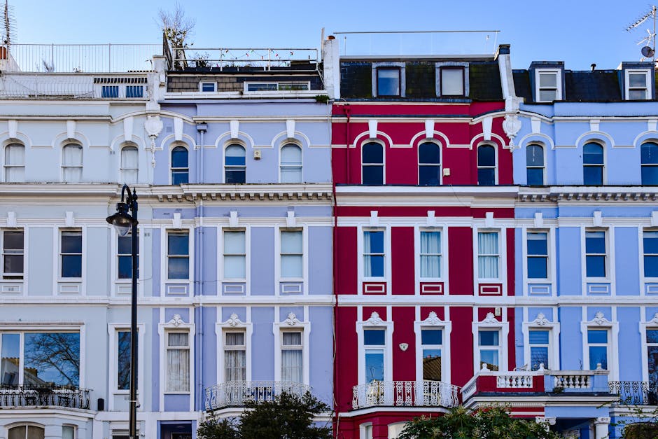 Photograph of a row of Victorian-style terrace houses on a clear day, featuring ornate white and red painted facades with multiple sash windows, decorative cornices, and iron balcony railings. In the foreground, a vintage-style street lamp is visible, and a moving company vehicle might be parked nearby, suggesting ongoing home relocation or furniture transport activities typical of house removals in Ladbroke Grove (W11). The image captures the external environment where a professional removals service like [COMPANY_NAME] could be involved in packing, loading, or transporting household items during a residential move, with the detailed architecture highlighting the characteristic features of Notting Hill's residential streets.