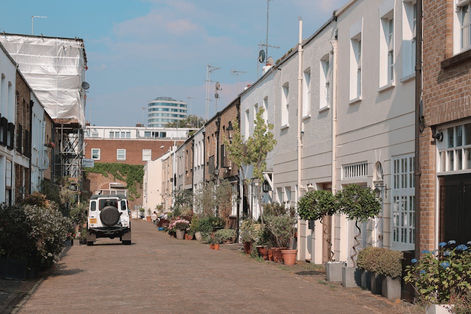 A narrow residential street in Notting Hill lined with terraced houses on both sides, featuring white and brick facades with large windows. In the foreground, a white van is parked on cobblestone paving, and nearby, there are potted plants and small trees placed along the pavement and outside the house entrances. The street is clear of traffic, with no people visible, and a modern, round office building can be seen in the background under a partly cloudy sky. The scene suggests a quiet home relocation or furniture transport process, with the van positioned for loading or unloading household items, possibly as part of a professional removal service by [COMPANY_NAME], supporting packing and moving activities essential for house removals in the Ladbroke Grove (W11) area.