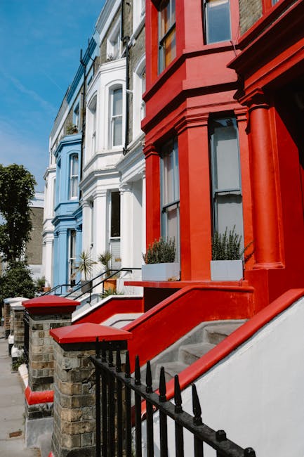 The image shows a row of Victorian-style residential buildings with colorful facades, including a prominent red-painted house with white stairs leading up to the entrance, which is partially visible. Adjacent to it are light blue and white houses with similar architectural features, such as bay windows and decorative moldings. The front steps are made of concrete, with some having black metal railings and stone pillars supporting small brick walls. Potted plants with green foliage are placed on the steps and window sills, adding some natural detail. The pavement in front of the houses is clean and well-maintained, and the sky above is clear, indicating daylight. This scene reflects an urban residential area suitable for professional house removals and furniture transport, aligning with services offered by companies like [COMPANY_NAME] for home relocation and packing moving processes. The image emphasizes the typical setting for loading and unloading items, with front entrances prepared for efficient removal tasks.