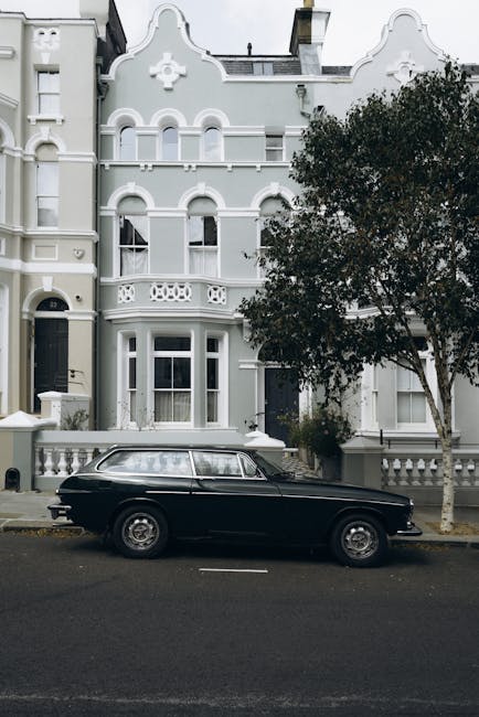 A black vintage station wagon parked on a residential street in front of a white Victorian-style townhouse featuring ornate architectural details, arched windows, decorative trim, and a small balcony. To the right of the vehicle, a large leafy tree partially obscures the building, and the street is lined with paving stones and curbside parking spaces. The scene is captured during daylight with diffused natural lighting, illustrating the typical urban environment for a home relocation or furniture transport process, with the vehicle likely used for moving belongings as part of local removals services near Notting Hill Gate, as described on the removal companies website.