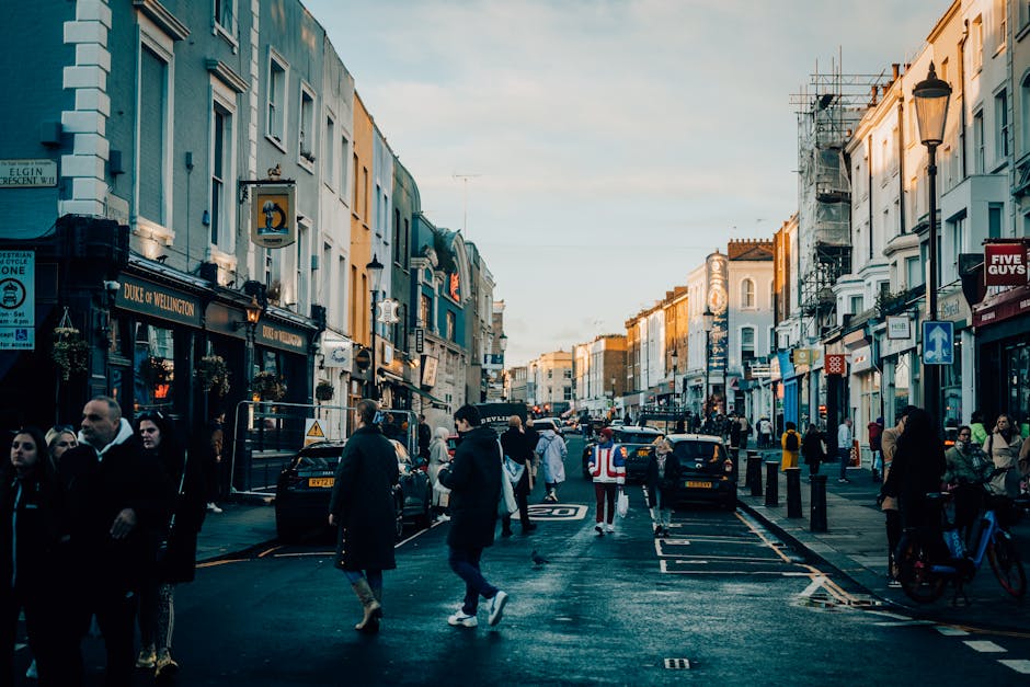 A busy urban street near Notting Hill Gate during daylight, with pedestrians walking on the pavement and crossing the road. Multiple black and yellow taxis are seen parked along the curb, alongside cars and delivery vehicles. The street is lined with multi-storey buildings housing cafes, shops, and offices, some with signs and shopfronts visible, including one with a blue sign and another with a red sign. Construction scaffolding and scaffolding netting are present on the upper floors of some buildings, indicating ongoing development or renovations. The sky above is partly cloudy with natural daylight illuminating the scene, capturing the typical vibrant atmosphere of a central London neighbourhood. This setting suggests the location for a home relocation or moving service, with the urban environment and street activity highlighting the logistical considerations for removals near Notting Hill, as managed by [COMPANY_NAME].