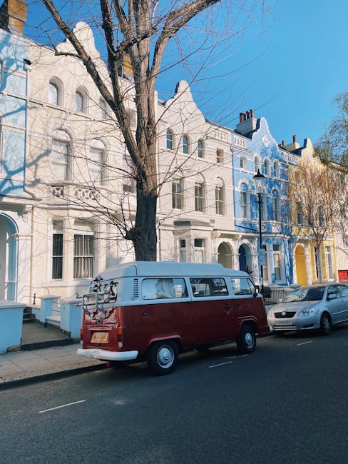 A black vintage station wagon parked on a residential street in front of a white Victorian-style townhouse featuring ornate architectural details, arched windows, decorative trim, and a small balcony. To the right of the vehicle, a large leafy tree partially obscures the building, and the street is lined with paving stones and curbside parking spaces. The scene is captured during daylight with diffused natural lighting, illustrating the typical urban environment for a home relocation or furniture transport process, with the vehicle likely used for moving belongings as part of local removals services near Notting Hill Gate, as described on the removal companies website.